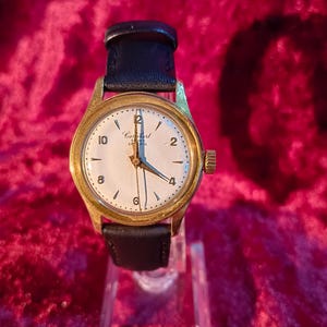 May include: A gold-toned analog watch with a white face and black leather band. The watch face displays the brand name "Combert" and features black numerals and hands. The watch is displayed on a clear stand against a red background.