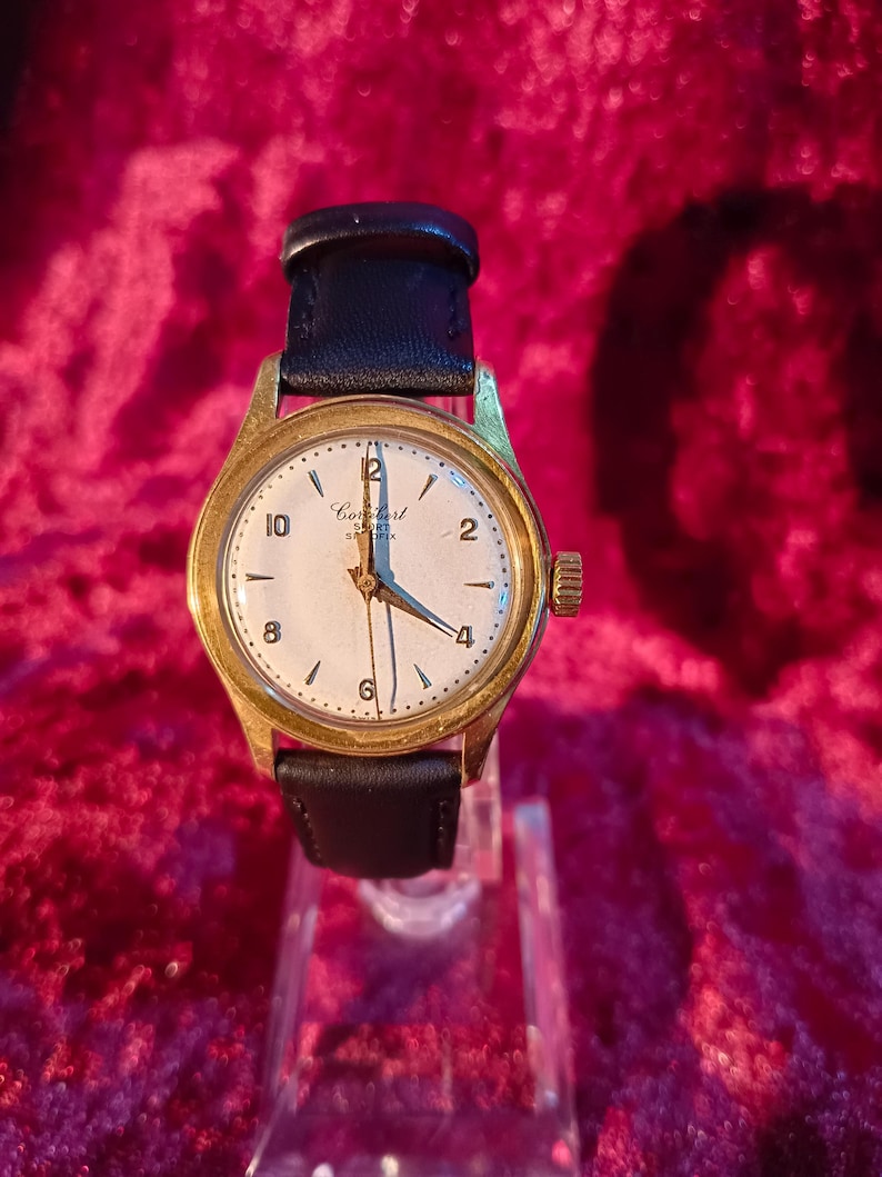 May include: A gold-toned analog watch with a white face and black leather band. The watch face displays the brand name "Combert" and features black numerals and hands. The watch is displayed on a clear stand against a red background.