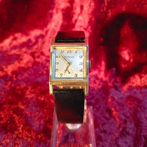 Op de afbeelding: Een vintage Wittnauer polshorloge met een rechthoekige goudkleurige kast en een zwarte leren band. De wijzerplaat van het horloge is crèmekleurig met zwarte cijfers en wijzers. Het horloge wordt tentoongesteld op een transparante standaard tegen een rode achtergrond.