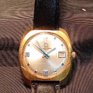May include: A gold-toned wristwatch with a black leather band. The watch face is silver with blue hands and blue markers at the hour positions. The word "UNICORN" is printed on the watch face. The date is displayed at the 3 o'clock position.