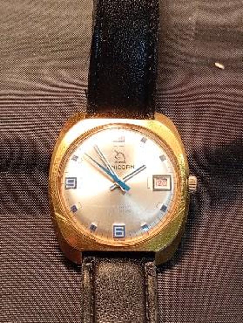 May include: A gold-toned wristwatch with a black leather band. The watch face is silver with blue hands and blue markers at the hour positions. The word "UNICORN" is printed on the watch face. The date is displayed at the 3 o'clock position.