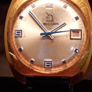 May include: A gold-toned vintage wristwatch with a light-colored face. The watch features a unicorn logo, blue hands, and a date display. The watch has a black band and a rectangular gold case.