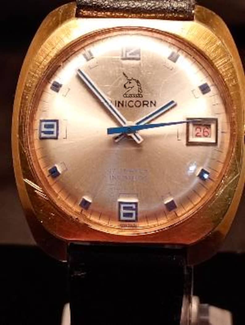 May include: A gold-toned vintage wristwatch with a light-colored face. The watch features a unicorn logo, blue hands, and a date display. The watch has a black band and a rectangular gold case.