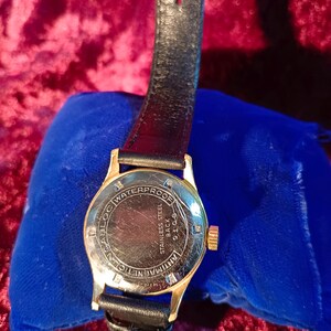 May include: A gold-toned wristwatch with a black leather band. The watch face is visible, displaying the words "WATERPROOF", "STAINLESS STEEL BACK", and "ANTIMAGNETIC". The watch is resting on a blue velvet surface.