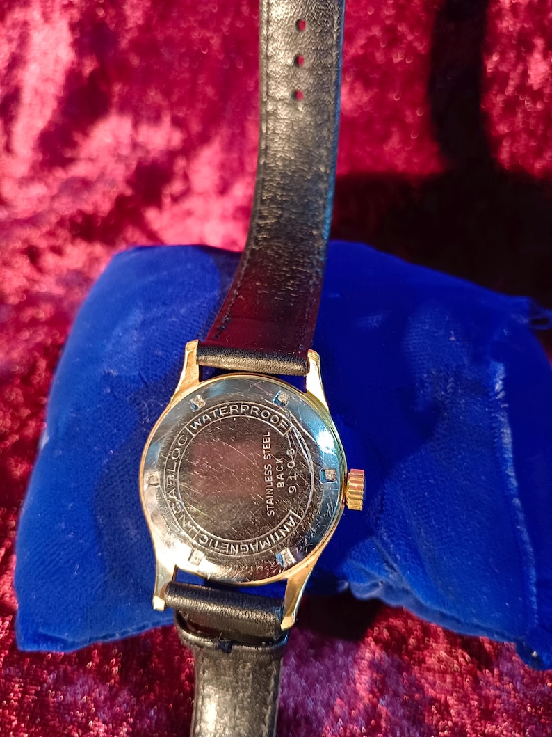May include: A gold-toned wristwatch with a black leather band. The watch face is visible, displaying the words "WATERPROOF", "STAINLESS STEEL BACK", and "ANTIMAGNETIC". The watch is resting on a blue velvet surface.