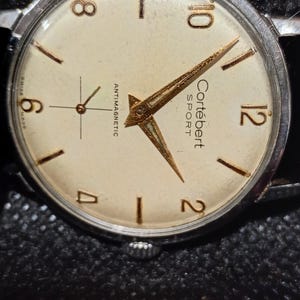 May include: Vintage Cortébert Sport wristwatch with a cream-colored face, gold-tone hands and markers, and a silver-tone case. The watch face displays the text "ANTIMAGNETIC" and "SWISS MADE". The watch has a black leather band.