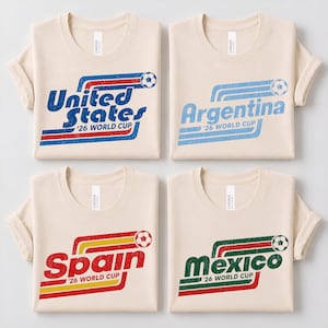May include: Four beige crew-neck t-shirts featuring distressed graphic prints of soccer balls and stripes. The shirts display the text United States, Argentina, Spain, and Mexico, each accompanied by the phrase '26 WORLD CUP in a retro-inspired design.