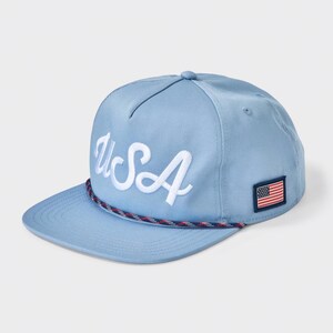 May include: A light blue baseball cap featuring the text USA embroidered in white script on the front panel. The cap includes a multi-colored braided rope detail across the base of the crown and a small rectangular American flag patch on the side.