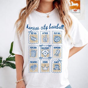 May include: White t-shirt with a blue and gold design featuring the text "Kansas City Baseball." The design includes illustrations of baseball-related items like a hot dog, popcorn, and a baseball field.