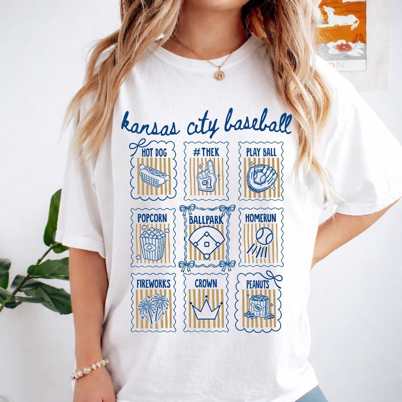 May include: White t-shirt with a blue and gold design featuring the text "Kansas City Baseball." The design includes illustrations of baseball-related items like a hot dog, popcorn, and a baseball field.
