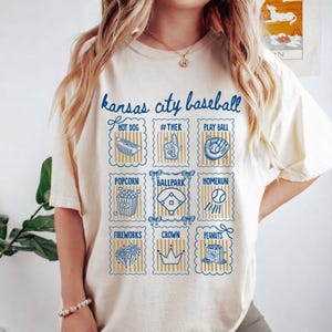 May include: Cream-colored t-shirt with a blue graphic design featuring the text "Kansas City Baseball." The design includes illustrations of baseball-related items like a hot dog, popcorn, and a baseball field. The shirt is made of a soft material.