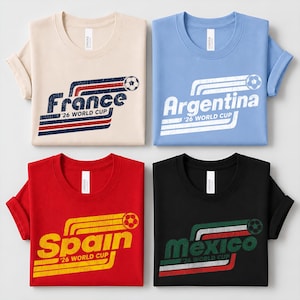 May include: Four t-shirts in beige, light blue, red, and black, each featuring a distressed graphic with a soccer ball and the text '26 WORLD CUP' alongside the country names France, Argentina, Spain, and Mexico in a retro striped design.