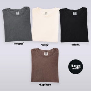 Puede incluir: Vista superior de cuatro camisetas dobladas en colores: pimienta, marfil, negro y espresso. Las camisetas son lisas, con cuello redondo y mangas cortas. El nombre de la marca "Luxe threads" se muestra en un c&iacute;rculo negro.