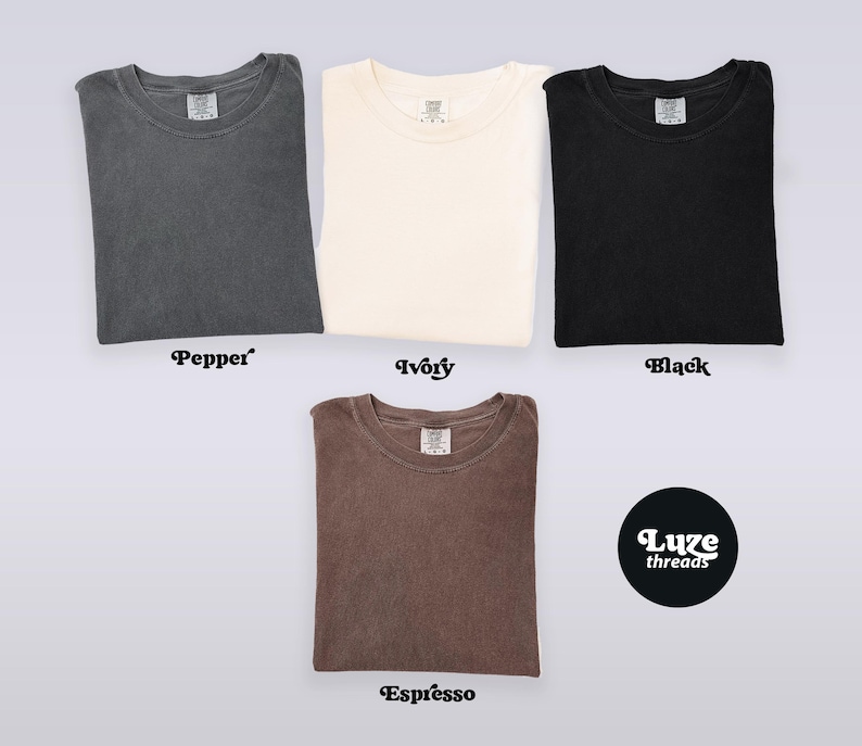Puede incluir: Vista superior de cuatro camisetas dobladas en colores: pimienta, marfil, negro y espresso. Las camisetas son lisas, con cuello redondo y mangas cortas. El nombre de la marca "Luxe threads" se muestra en un c&iacute;rculo negro.