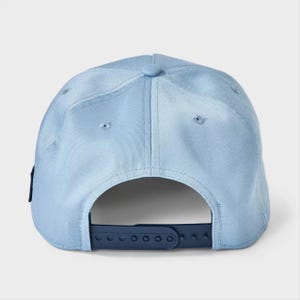 May include: A light blue baseball cap featuring a structured crown with embroidered eyelets and a dark blue plastic snapback closure at the rear. The cap is constructed from a solid-colored fabric and includes a small dark blue tab on the side.