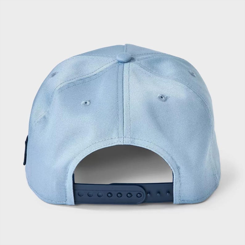 May include: A light blue baseball cap featuring a structured crown with embroidered eyelets and a dark blue plastic snapback closure at the rear. The cap is constructed from a solid-colored fabric and includes a small dark blue tab on the side.