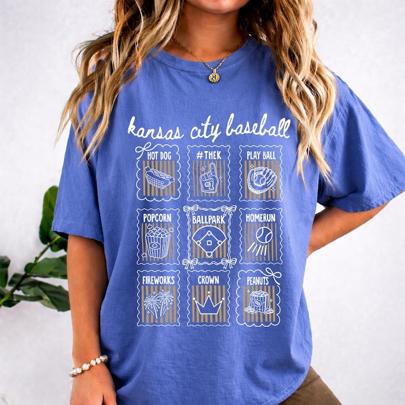 May include: A blue t-shirt with the words "Kansas City Baseball" in white script. Below the text are nine white and gold illustrations of baseball-related items, including a hot dog, popcorn, and a baseball.