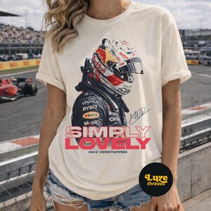 Puede incluir: Camiseta color crema con un dise&ntilde;o de un casco de carreras, el logotipo de Red Bull y el texto "SIMPLY LOVELY" en rosa y rojo. La camiseta tambi&eacute;n incluye el nombre "MAX VERSTAPPEN" debajo del texto.