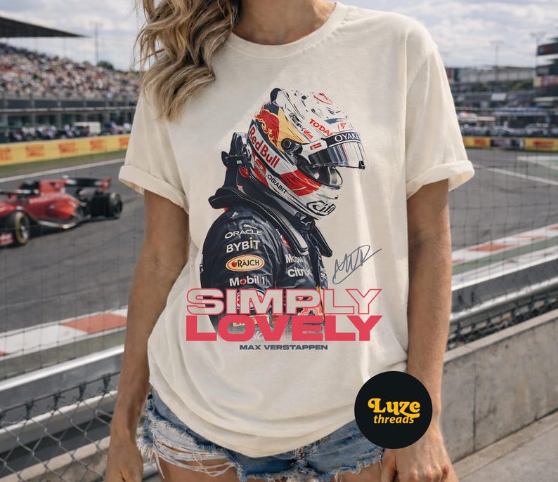 Puede incluir: Camiseta color crema con un dise&ntilde;o de un casco de carreras, el logotipo de Red Bull y el texto "SIMPLY LOVELY" en rosa y rojo. La camiseta tambi&eacute;n incluye el nombre "MAX VERSTAPPEN" debajo del texto.