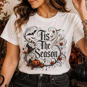 May include: A white t-shirt featuring a graphic print with the text 'Tis the Season' surrounded by illustrations of a jack-o'-lantern, a witch hat, a ghost, autumn leaves, and spiderwebs, all framed by an ornate black decorative border.