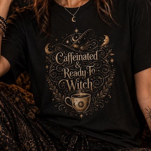 Op de afbeelding: Een zwart t-shirt met een gouden grafische print van een heksenhoed, een koffiekopje en de tekst Caffeinated & Ready To Witch, omringd door sierlijke, wervelende botanische patronen, sterren en maansikkels op de voorzijde.
