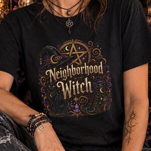 May include: A black short-sleeved t-shirt featuring a graphic print with the text Neighborhood Witch. The design includes a witch hat, a pentagram, a black cat, and a boot in shades of gold and purple, set against a dark, ornate background.