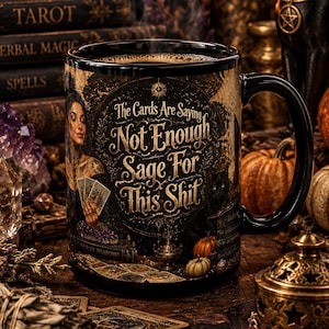 Può includere: Una tazza in ceramica nera con un'illustrazione grafica di una persona che tiene in mano carte dei tarocchi, circondata da immagini mistiche e dal testo, The Cards Are Saying Not Enough Sage For This Shit, con finitura lucida e manico.