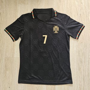 Może przedstawiać: Czarna koszulka piłkarska z numerem 7 w kolorze złotym. Koszulka ma powtarzający się wzór herbów i złoty emblemat. Kołnierz i mankiety rękawów mają zielone, czerwone i żółte paski.