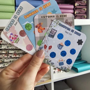 May include: Four square scratch-off cards representing the seasons. The cards feature the text Spring Is Here, Summer Is Here, Autumn Is Here, and Winter Is Here, each with Save £20 printed below. They include seasonal illustrations and circular scratch-off areas.