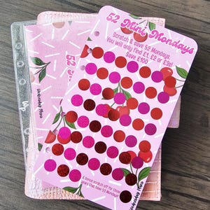 May include: A pink savings tracker card titled 52 Mini Mondays with cherry illustrations and rows of scratch-off circles. The text reads: Scratch & save 52 Mondays. You will only find £1, £2 or £3s! Save £100. A bonus scratch off for those years that have 53 Mondays.