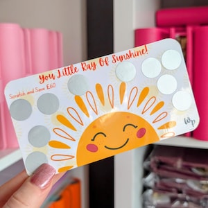 May include: A white card featuring a smiling sun illustration and the text "You Little Ray Of Sunshine!". The card has silver scratch-off circles and the text "Scratch and Save". The sun is orange and yellow with a smiling face.