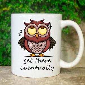 Könnte beinhalten: Eine weiße Keramik-Kaffeetasse mit einer Cartoon-Illustration einer braunen Eule mit gelben Augen, geschlossenen Augenlidern und einem schläfrigen Ausdruck. Die Eule trägt ein pinkfarbenes und weiß gemustertes Hemd. Der Text "get there eventually" ist unter der Eule aufgedruckt.