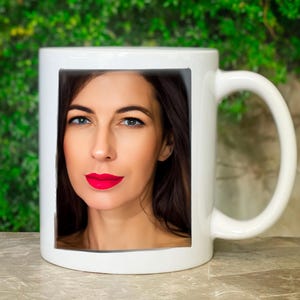 Könnte beinhalten: Weiße Keramiktasse mit einem Foto einer Frau mit langen dunklen Haaren und roten Lippen. Die Frau schaut direkt in die Kamera.