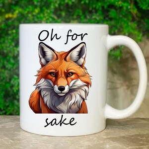 Könnte beinhalten: Weiße Keramiktasse mit einer Illustration eines Fuchses in Orange- und Weißtönen. Der Becher ist mit den Worten "Oh for sake" in Schwarz bedruckt. Die Tasse hat einen gebogenen Griff.
