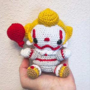 Puede incluir: Muñeca de payaso de ganchillo con un globo rojo. El payaso tiene una cara blanca con detalles rojos, ojos negros y pelo amarillo. Viste un traje gris con ribetes rojos y zapatos marrones y amarillos. El globo rojo está en su mano izquierda.