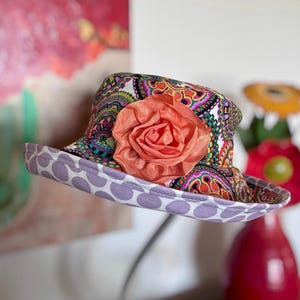 May include: A colorful paisley print hat with a wide brim featuring a large peach-colored fabric rose. The brim is lined with a lavender fabric with white polka dots. A stylish accessory.