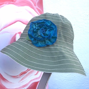 Puede incluir: Un sombrero de sol a rayas verde y blanco con una gran flor de tela azul en la parte delantera.