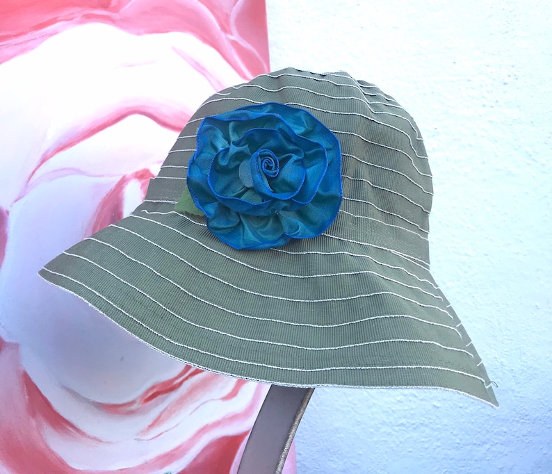 Puede incluir: Un sombrero de sol a rayas verde y blanco con una gran flor de tela azul en la parte delantera.
