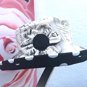 May include: A bucket hat featuring a cream-colored crown with black vintage-style script and postal stamp graphics. The hat is adorned with a fabric flower with cream petals and a black center, finished with a black brim featuring white polka dots.