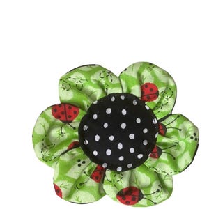 May include: A fabric flower with six green petals featuring a ladybug and leaf pattern. The center is a black circle with white polka dots. The flower is a craft item.
