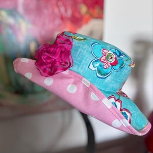 May include: A colorful sun visor with a pink polka dot brim and a turquoise top adorned with floral patterns. A vibrant fuchsia ribbon adds a decorative touch. This accessory is perfect for sunny days.