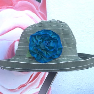 Puede incluir: Un sombrero de sol a rayas verde y blanco con una gran rosa de tela azul en la parte delantera.