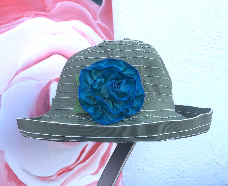 Puede incluir: Un sombrero de sol a rayas verde y blanco con una gran rosa de tela azul en la parte delantera.