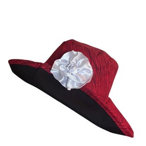 May include: A wide-brimmed hat featuring a red fabric crown with a black wood grain pattern and a solid black brim. A large, white, multi-layered satin fabric flower is attached to the front of the crown, providing a distinct contrast to the red.
