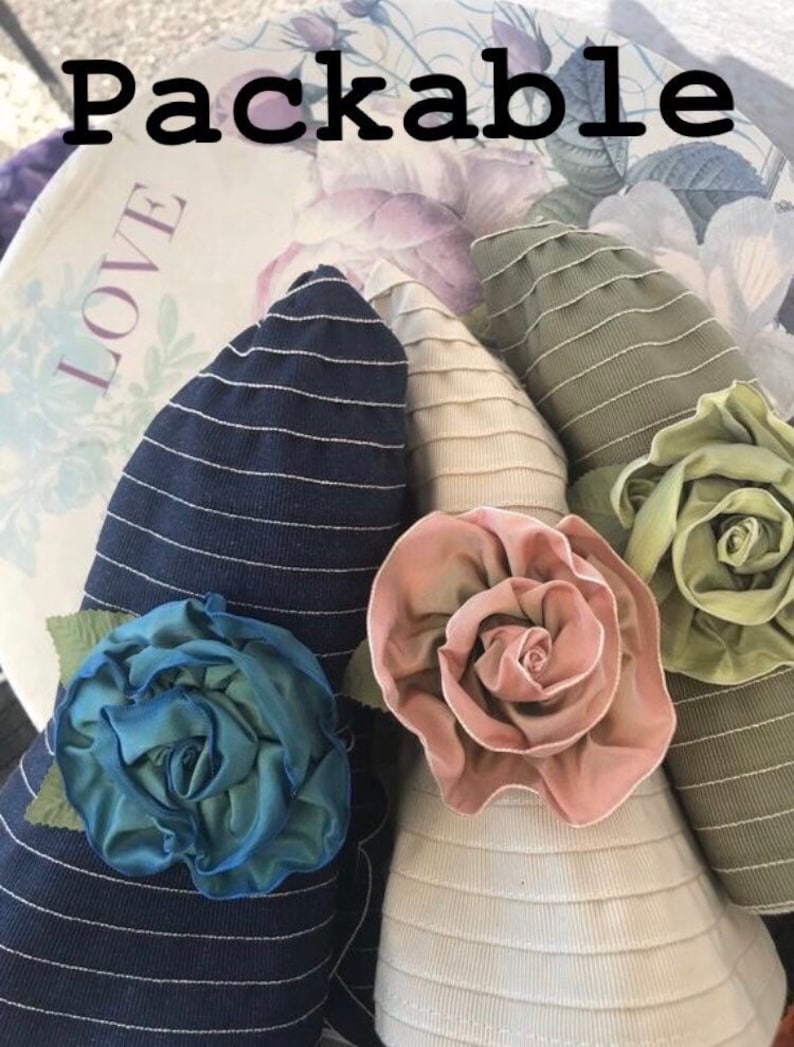 Puede incluir: Tres sombreros de sol plegables con rosetas de tela de diferentes colores. Los sombreros son a rayas y tienen un ala ancha. Las rosetas son en tonos de rosa, verde y azul.
