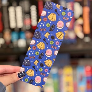 May include: A rectangular bookmark with a blue background featuring a repeating pattern of yellow raincoats, black cats, pink cakes, green and pink buttons, black keys, dragonflies, and dark blue sweaters with white stars scattered throughout.
