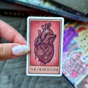 May include: A rectangular sticker featuring a stylized illustration of a human heart with blood dripping from it. The heart is a deep red color with glittery accents. The text "THE HEARTEATER" is printed at the bottom of the sticker.