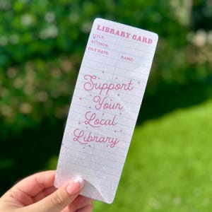 May include: A white library card with the text "Support Your Local Library" in pink script. The card has lines for title, author, due date, and name. The background is a blurred green.