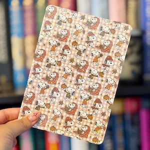 May include: A rectangular e-reader skin featuring a repeating pattern of Snoopy characters in autumn attire, including scarves and hats, holding pumpkins or wrapped in blankets. The design is set against a light brown gingham background with holographic stars.