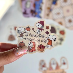 Può includere: Un adesivo fustellato che raffigura Snoopy in varie pose a tema autunnale, tra cui tenere in mano una zucca, indossare una sciarpa e avvolto in una coperta a quadri. Il testo "I love fall most of all" è scritto in corsivo nero al centro.